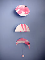 Moon phase shapes painted with pink and black ink stitched to create a wall hanging displayed on a blue background 