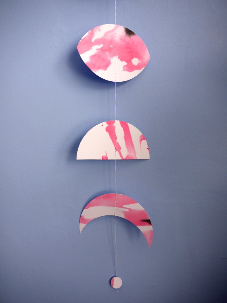 Moon phase shapes painted with pink and black ink stitched to create a wall hanging displayed on a blue background 