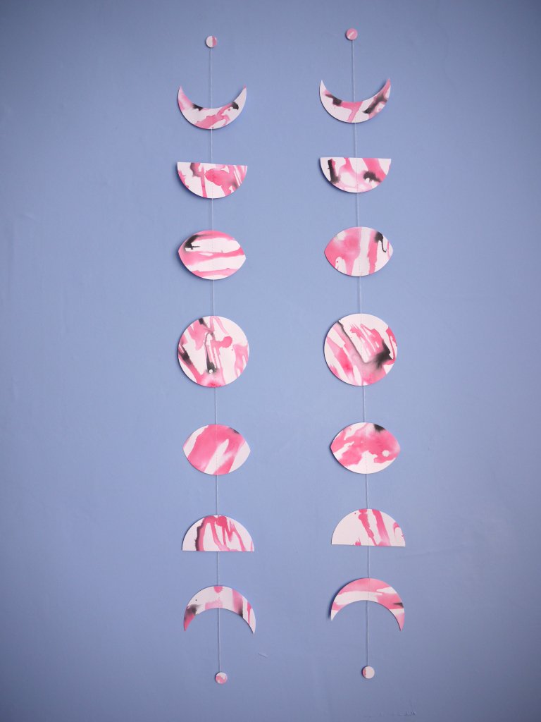 Two moon phase garlands painted slightly differently with pink and black ink stitched to create a wall hanging displayed on a blue background 