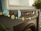 Paper garlands from Paper Street Dolls Ragnar collection. At a fireplace sits a multishape garland in cool tones, below a marbled circle garland 