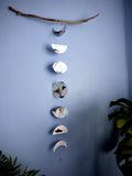 Moon phases wall hanging stitched paper garland painted with black and blue inks hung on driftwood on a blue wall with plants either side