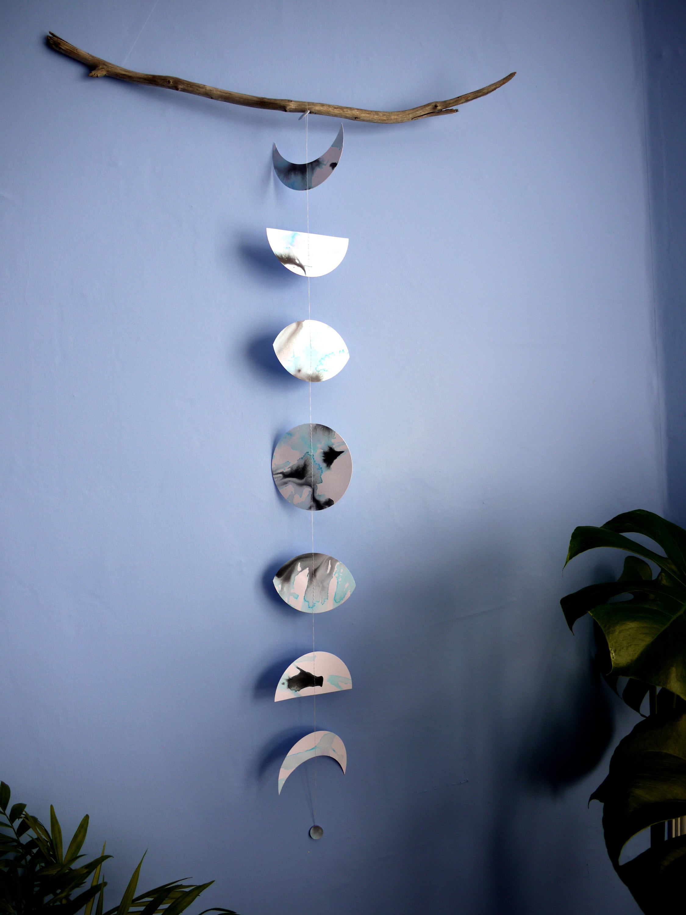 Moon phases wall hanging stitched paper garland painted with black and blue inks hung on driftwood on a blue wall with plants either side