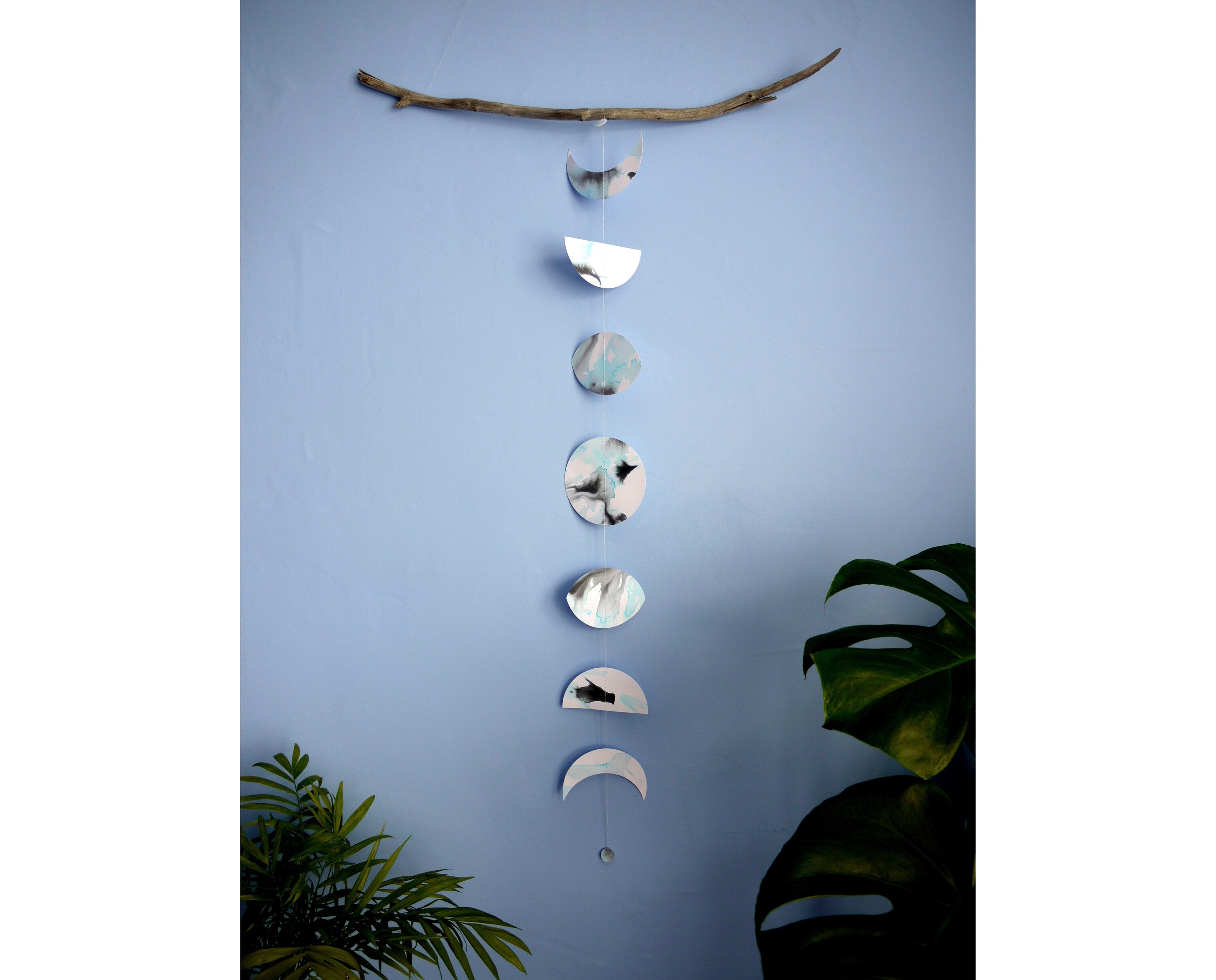 Moon phases wall hanging stitched paper garland painted with black and blue inks hung on driftwood on a blue wall with plants either side