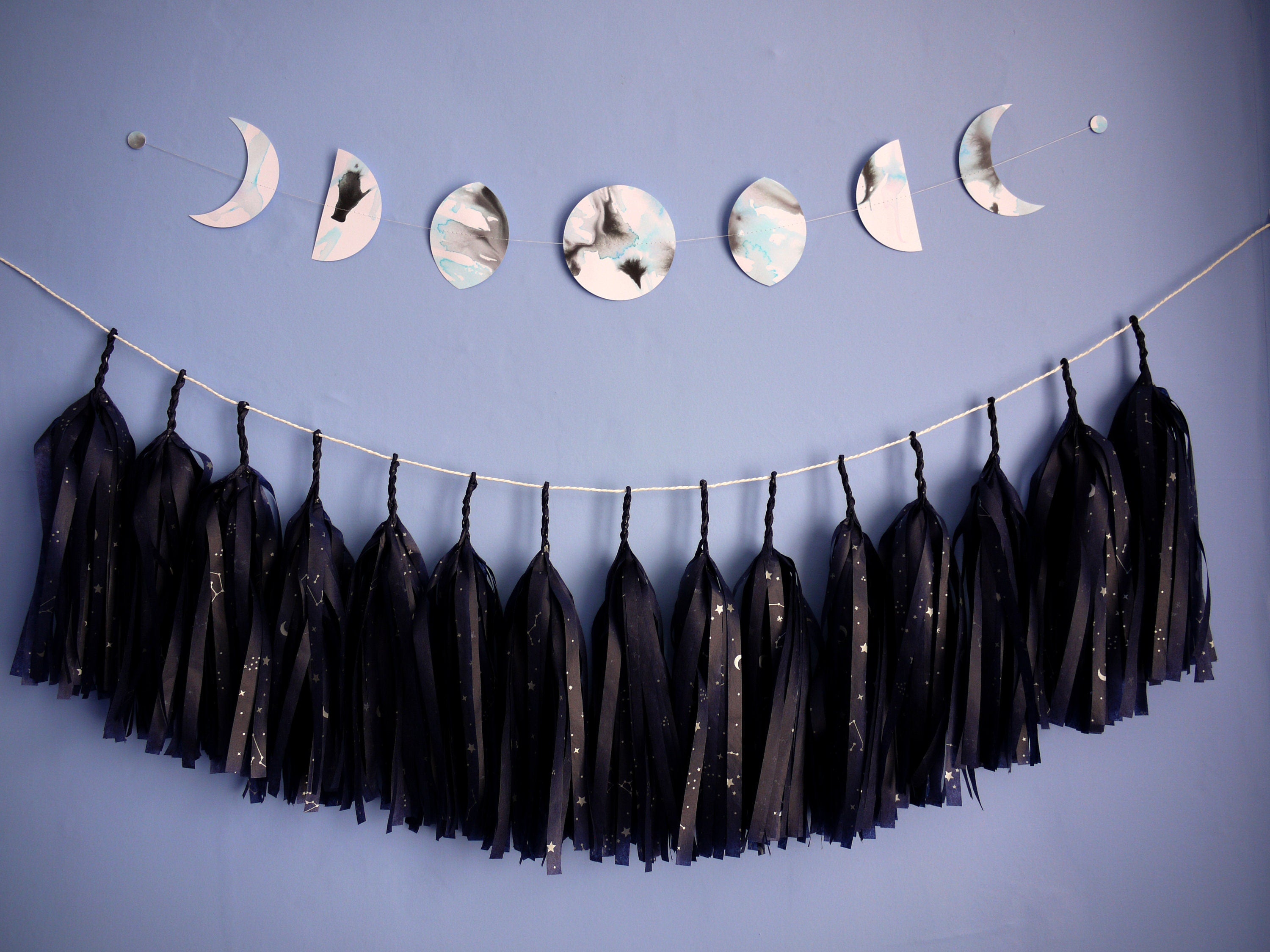 Moon phases wall hanging stitched paper garland painted with black and blue inks hung horizontally on a blue wall with a strand of navy blue tissue tassels hanging underneath 