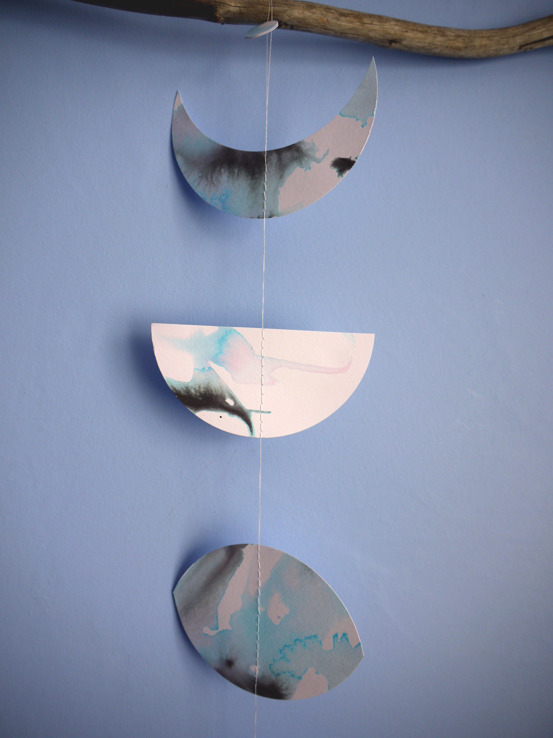 Moon phases wall hanging stitched paper garland painted with black and blue inks hung on driftwood on a blue wall