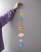 Colorful solar system model hanging on a gray wall with a person's arm visible.