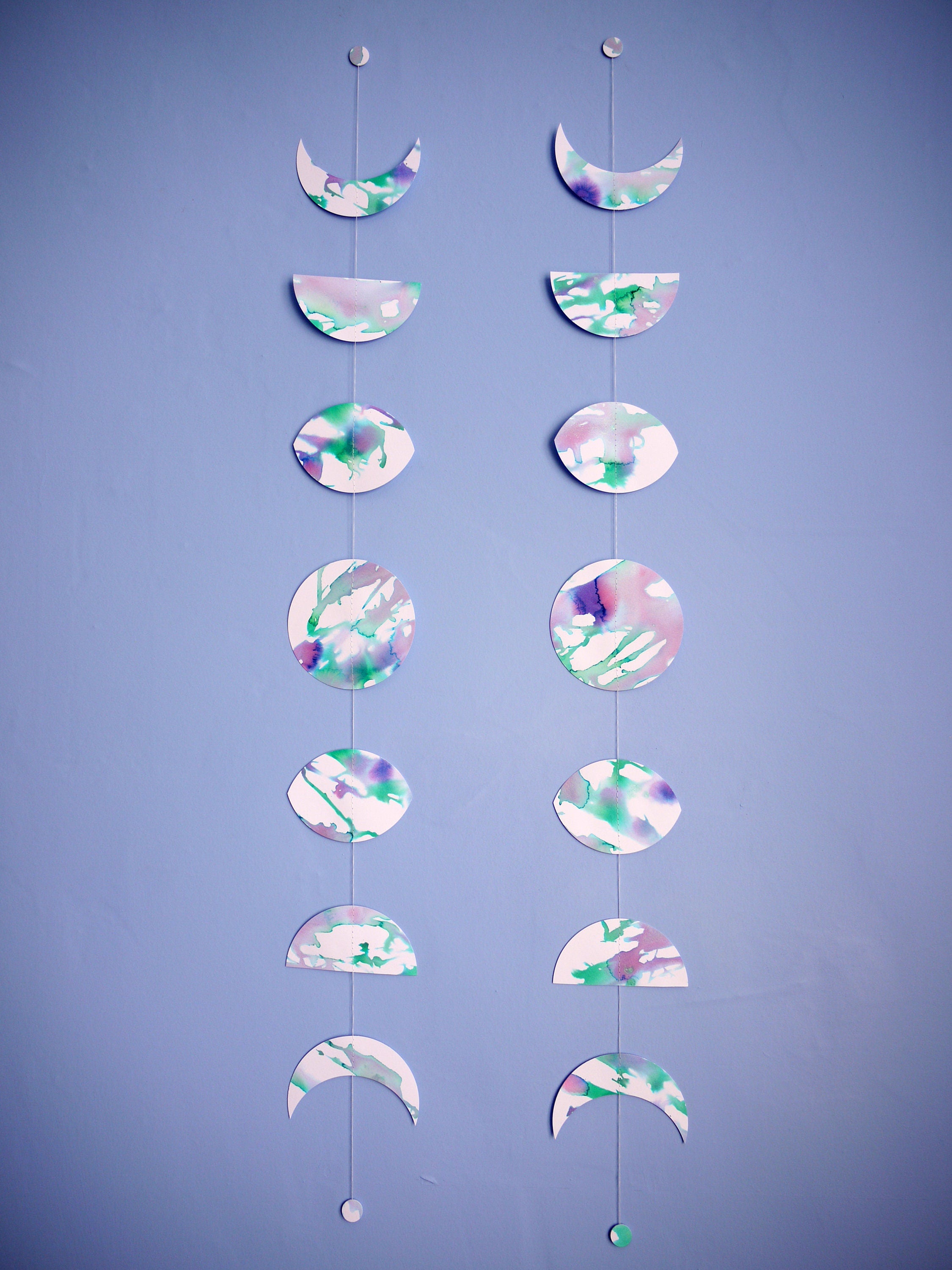 Two vertical moon phase shapes painted with green and purple pearlised ink stitched to create a wall garland displayed on a blue background