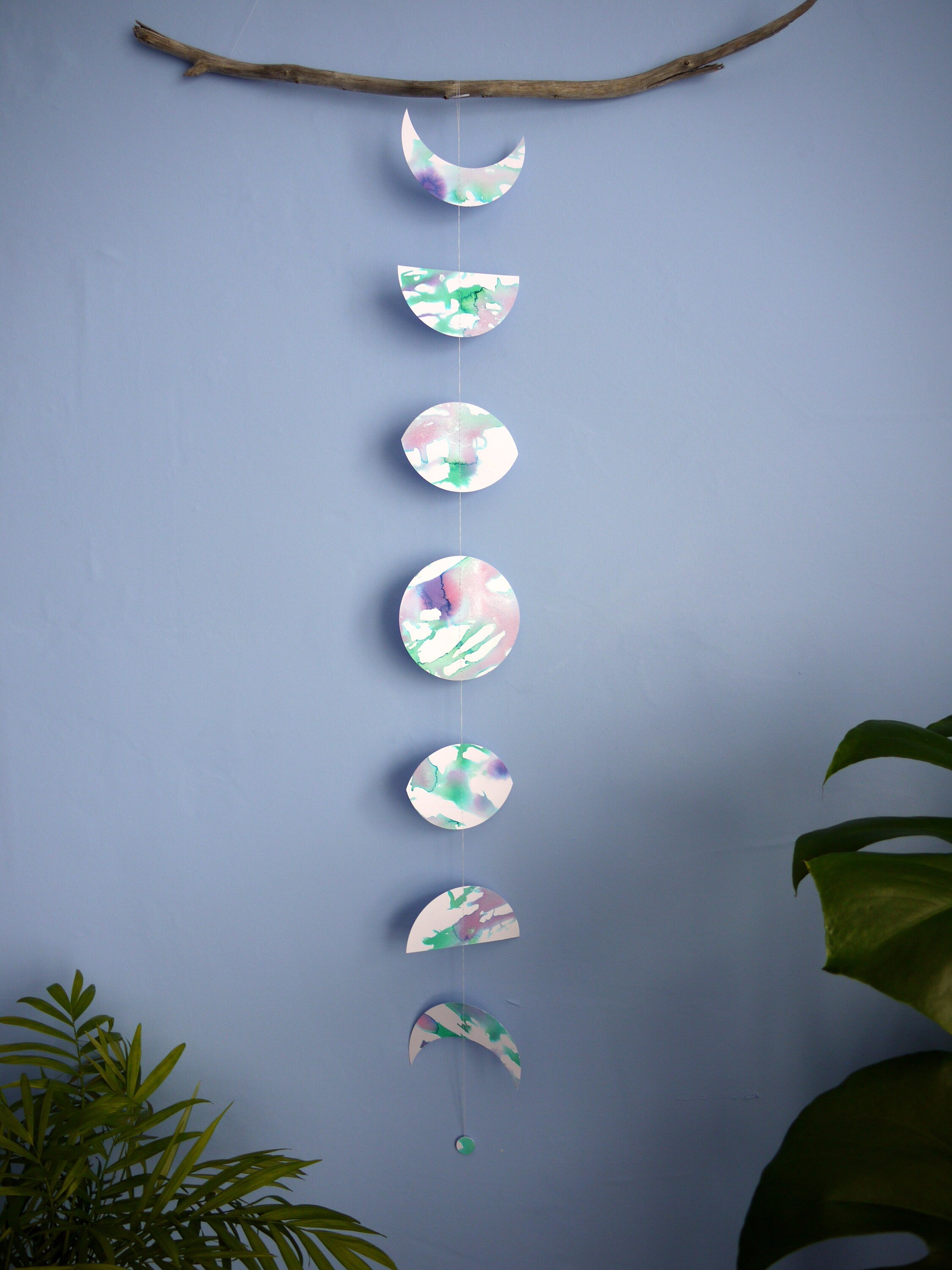 Vertical moon phase stitched paper garland painted with green and purple pearlised ink displayed as a wall hanging on a piece of driftwood displayed on a blue background with plants at either side
