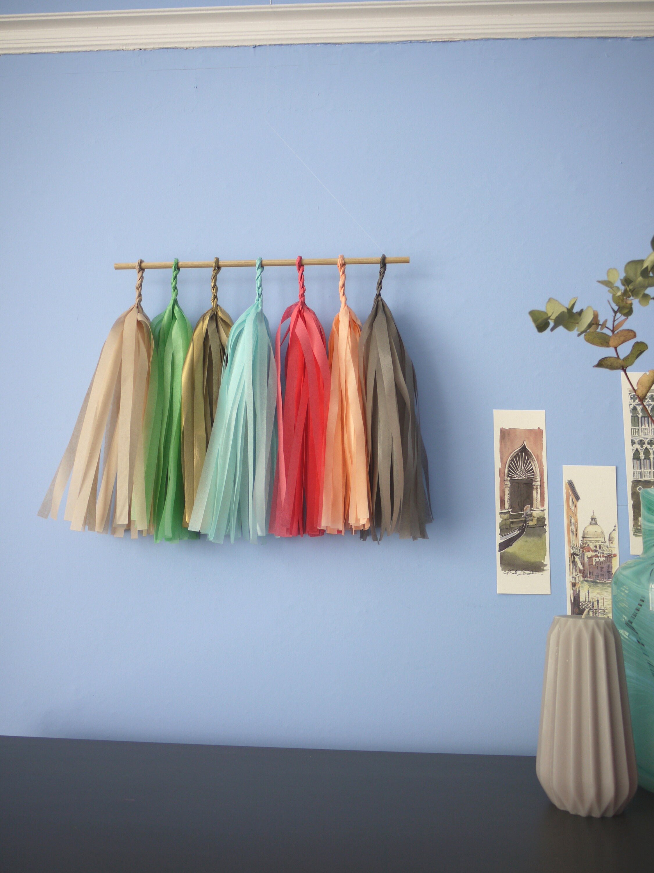 Soft coloured tassel wall hanging on a blue wall beside some Venice illustrations and trinkets