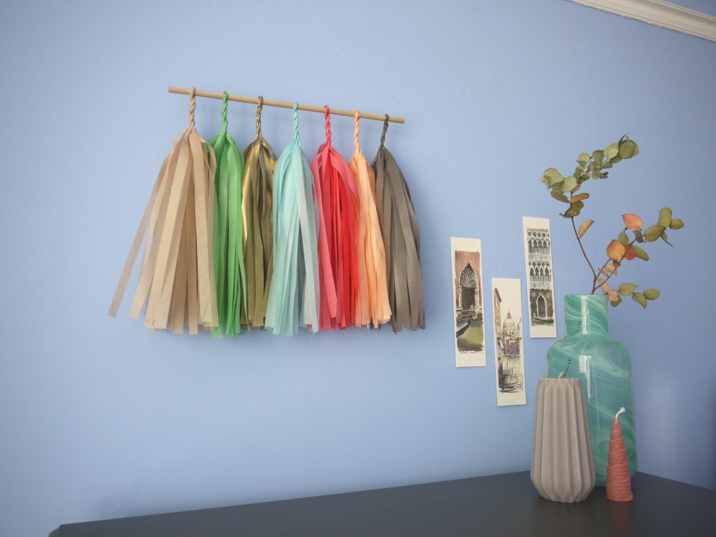 Soft coloured tassel wall hanging on a blue wall beside some Venice illustrations and trinkets