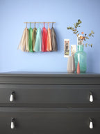 Soft coloured tassel wall hanging on a blue wall beside some Venice illustrations and trinkets on a chest of drawers