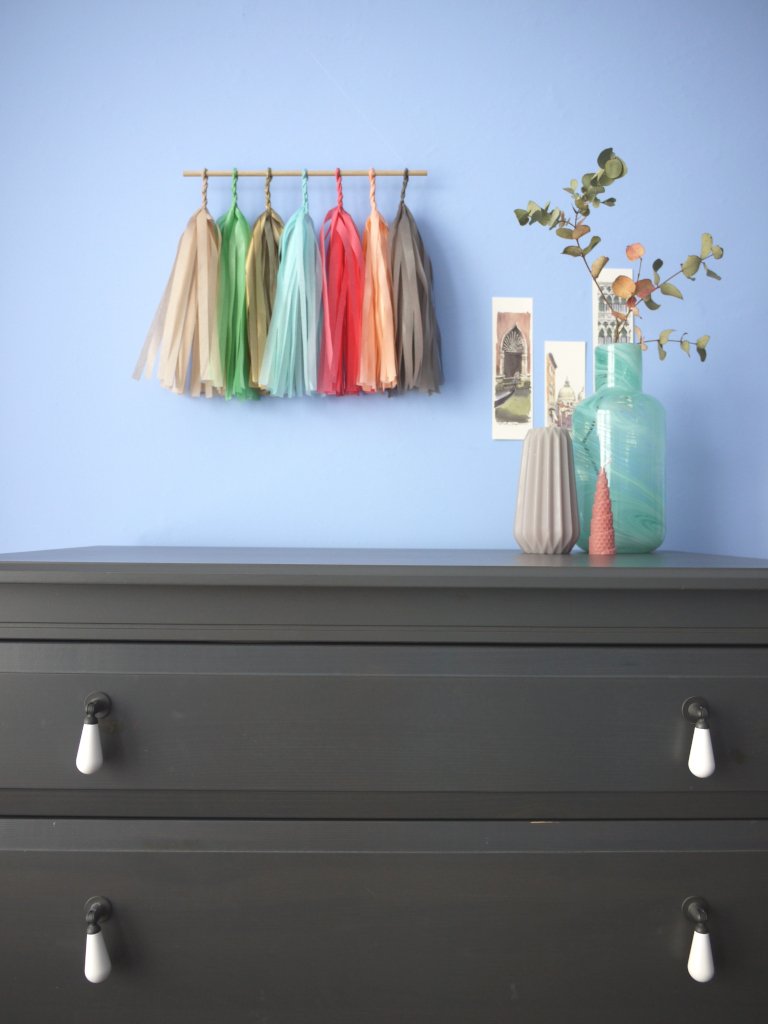 Soft coloured tassel wall hanging on a blue wall beside some Venice illustrations and trinkets on a chest of drawers
