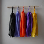 Blessed Samhain tassel wall hanging. Five colorful paper tassel garlands in black, cornflower, orange, purple, and yellow hang from a wooden dowel against a plain light gray wall.