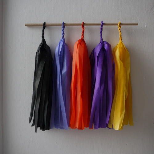 Blessed Samhain tassel wall hanging. Five colorful paper tassel garlands in black, cornflower, orange, purple, and yellow hang from a wooden dowel against a plain light gray wall.