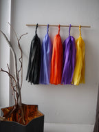 Blessed Samhain tassel wall hanging.  Five colorful paper tassel garlands in black, cornflower, orange, purple, and yellow hang from a wooden dowel on a white wall, with a decorative branch holder in the lower left corner.