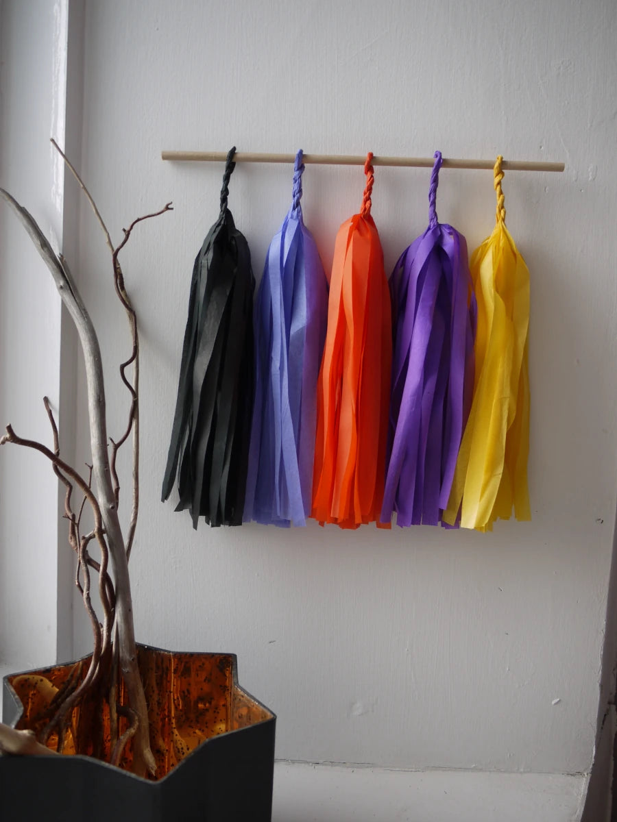 Blessed Samhain tassel wall hanging.  Five colorful paper tassel garlands in black, cornflower, orange, purple, and yellow hang from a wooden dowel on a white wall, with a decorative branch holder in the lower left corner.