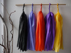 Blessed Samhain tassel wall hanging. 
Five colorful paper tassel garlands in black, cornflower, orange, purple, and yellow hang from a wooden dowel on a white wall, with a decorative branch holder in the lower left corner.