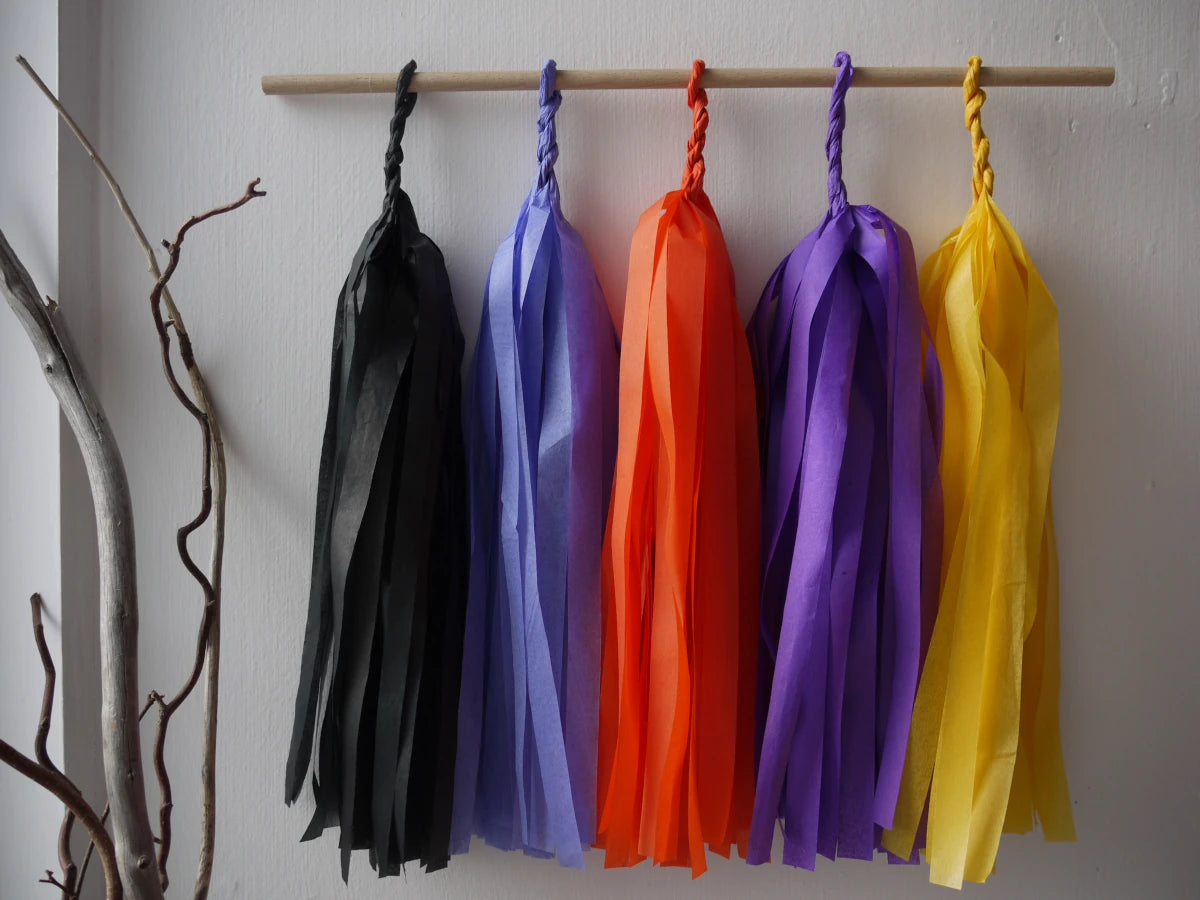 Blessed Samhain tassel wall hanging. 
Five colorful paper tassel garlands in black, cornflower, orange, purple, and yellow hang from a wooden dowel on a white wall, with a decorative branch holder in the lower left corner.