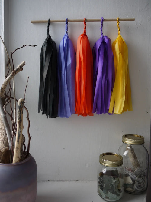 Blessed Samhain tassel wall hanging. 
Five colorful paper tassel in black, cornflower, orange, purple, and yellow hang from a wooden dowel against a white wall, with a ceramic vase of branches and two glass jars on a shelf below.