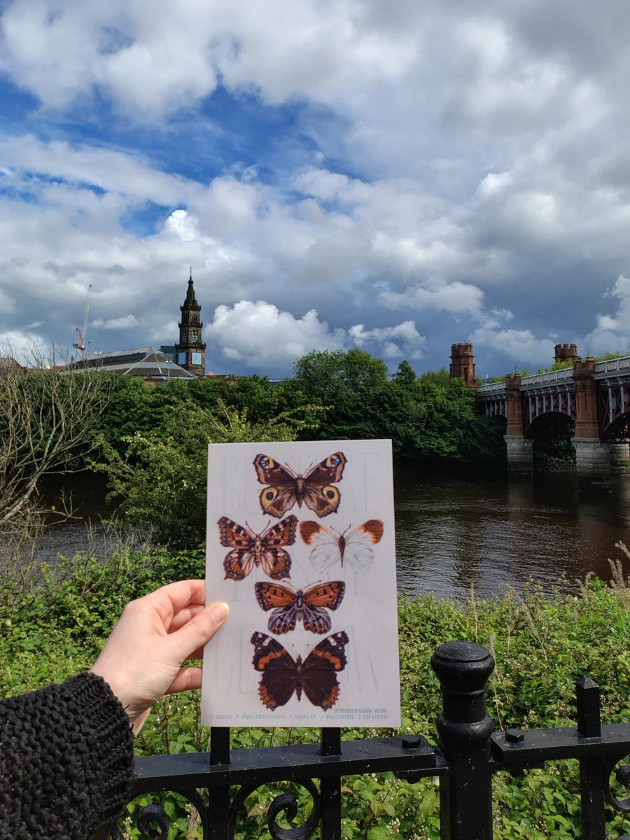 A hand holding a Butterfly riso print featuring butterflies of Glasgow, in front of the River Clyde.