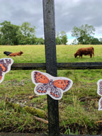 An illustrated small copper riso print butterfly shape from a butterfly garland. Naturalist home decor hanging outdoors in a grassy field with a fence and distant highland cows.