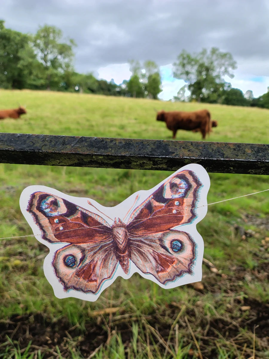 An illustrated peacock riso print butterfly shape from a butterfly garland. Naturalist home decor hanging outdoors in a grassy field with a fence and distant highland cows.