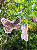 Two riso print butterfly shapes from a butterfly garland hanging from a buddleja tree branch in a lush green garden.