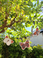 Three illustrated riso print butterfly shapes from a Paper Street Dolls butterfly garland, hanging from a budleja tree in a lush garden.