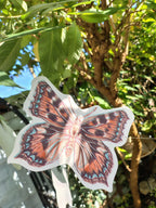 A riso print small tortoiseshell butterfly shape from a butterfly garland. Naturalist home decor photographed outdoors among buddleja tree branches and greenery.