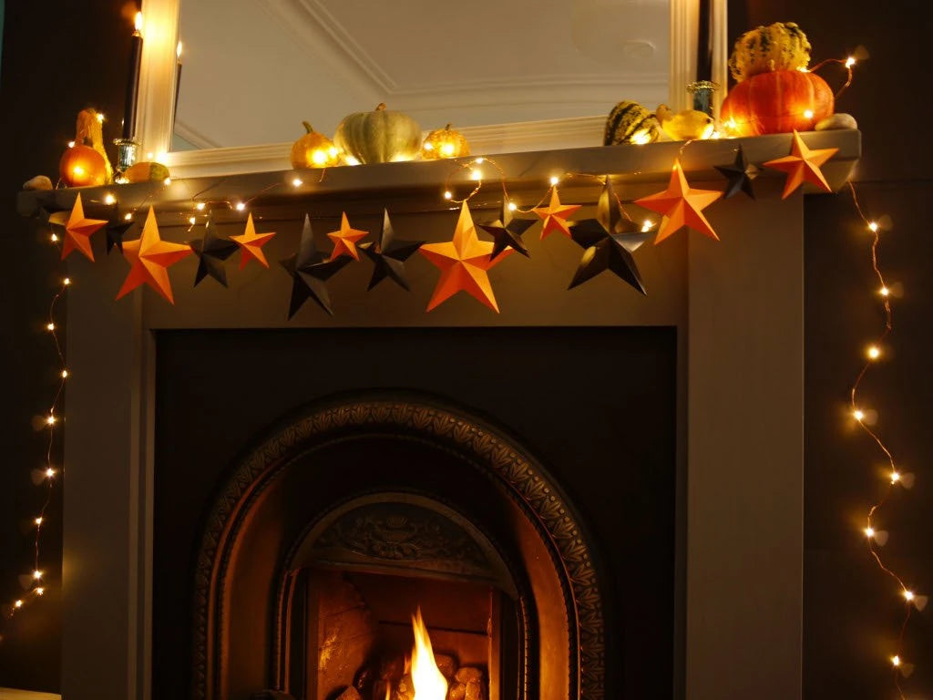 A cozy fireplace mantel decorated with hanging orange and black paper star garlands, warm string lights, small pumpkins, and gourds, with a fire burning below. Whimsigoth fun easy halloween crafts.
