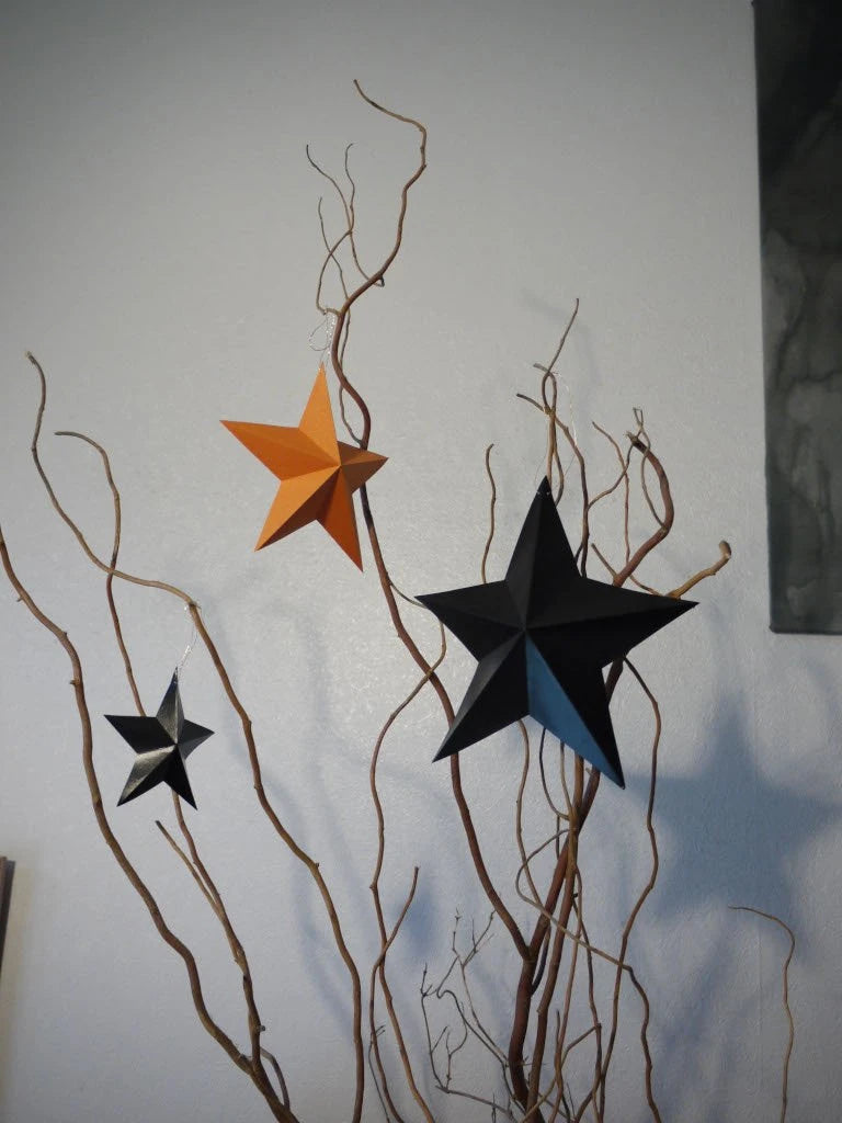 Three paper star ornaments in black, orange, hanging from bare brown branches against a plain light wall. Fun easy halloween crafts with the whimsigoth DIY paper star garland kit.