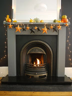 A gray fireplace mantel decorated with a whimsigoth paper garland of orange and black 3D stars, small pumpkins, and warm string lights, with a lit fire burning in the arched fireplace opening. Fun easy halloween crafts.