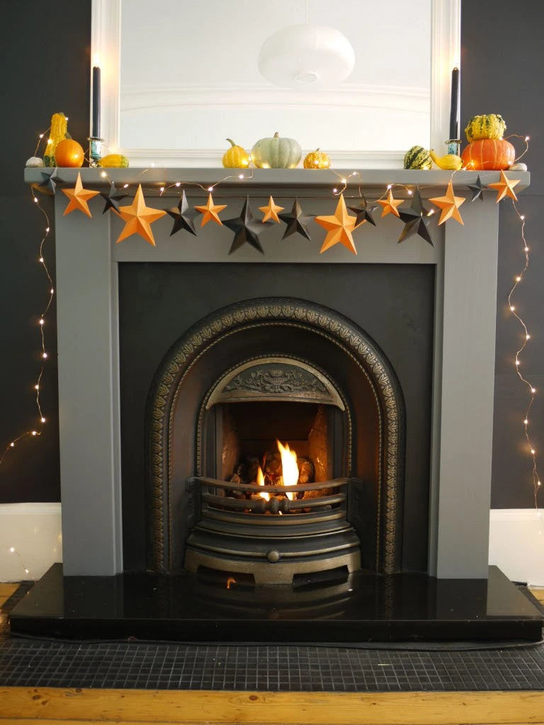 A gray fireplace mantel decorated with a whimsigoth paper garland of orange and black 3D stars, small pumpkins, and warm string lights, with a lit fire burning in the arched fireplace opening. Fun easy halloween crafts.