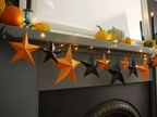 Fun easy halloween crafts with a DIY paper star garland kit. Orange and black 3D paper star garland hanging across a gray fireplace mantel decorated with pumpkins and warm string lights for a whimsigoth aesthetic