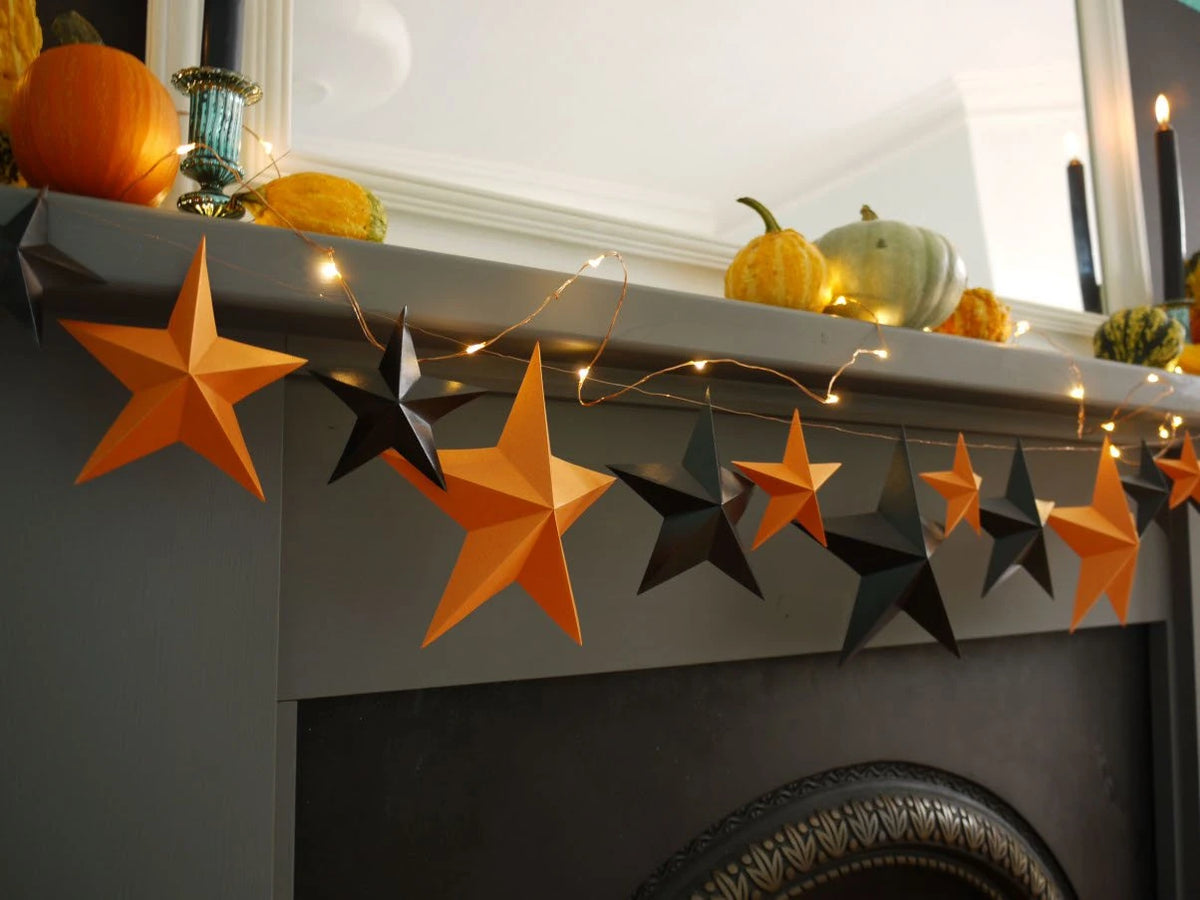 Fun easy halloween crafts with a DIY paper star garland kit. Orange and black 3D paper star garland hanging across a gray fireplace mantel decorated with pumpkins and warm string lights for a whimsigoth aesthetic