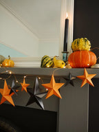 Whimsigoth orange and black Halloween paper star garland hanging across a gray fireplace mantel, styled with small pumpkins, a yellow gourd, a black candle, and warm string lights. Fun easy halloween crafts.
