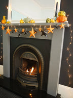 Whimsigoth Halloween fireplace decoration featuring a gray mantel with pumpkins, a DIY paper star garland of black and orange 3D stars cutouts, and warm fairy lights against a dark wall. Fun easy halloween crafts.