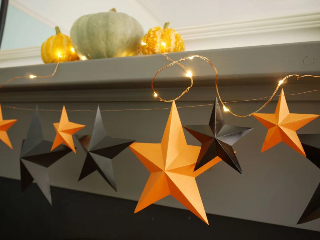 Fun easy halloween crafts with DIY paper star garland. Halloween decorations hanging on a mantel, featuring orange and black 3D paper stars with warm string lights, with decorative pumpkins in the background for full whimsigoth effect.