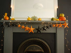 Whimsigoth Halloween fireplace mantel decorated with orange and black paper star garland, small pumpkins, string lights, and candles against a dark wall. Fun easy halloween crafts.