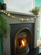 Decorative gold sewn paper garland draped at a fireplace surrounded by houseplants.