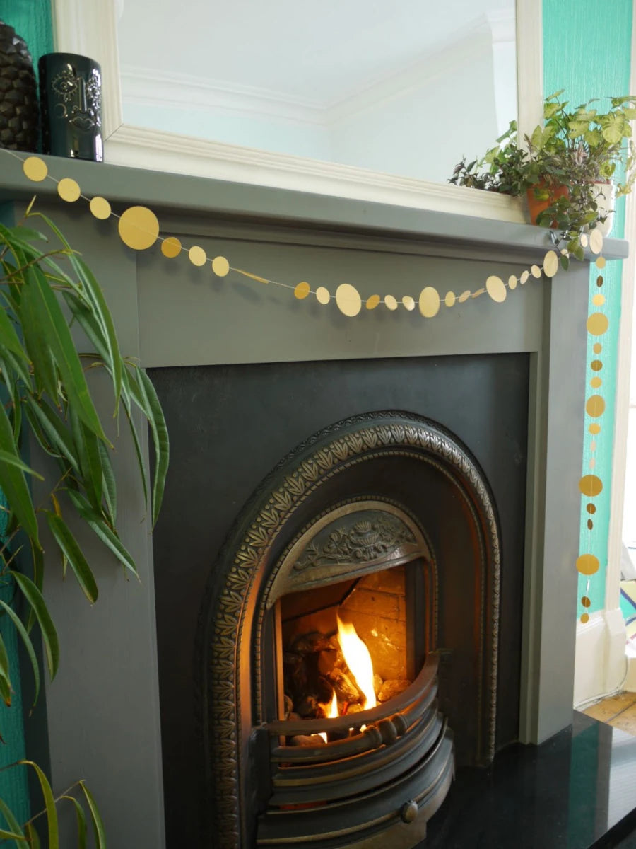 Decorative gold sewn paper garland draped at a fireplace surrounded by houseplants.