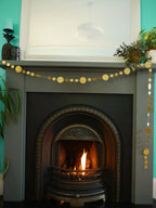 Decorative fireplace with a fire burning, gold circular paper garland, and plants against a teal wall.