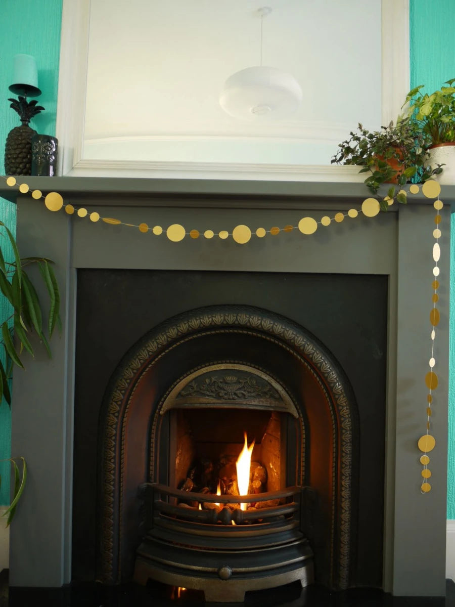 Decorative fireplace with a fire burning, gold circular paper garland, and plants against a teal wall.