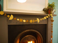 A gold geometric paper garland made of circles, squares, and triangles hangs across a dark fireplace mantel, with a mirror and potted plants above.