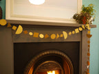 A gold geometric paper garland made of circles, squares, and triangles hangs across a dark fireplace mantel, with a mirror and potted plants above.