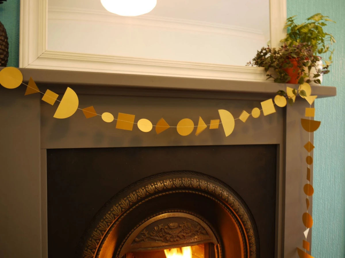 A gold geometric paper garland made of circles, squares, and triangles hangs across a dark fireplace mantel, with a mirror and potted plants above.