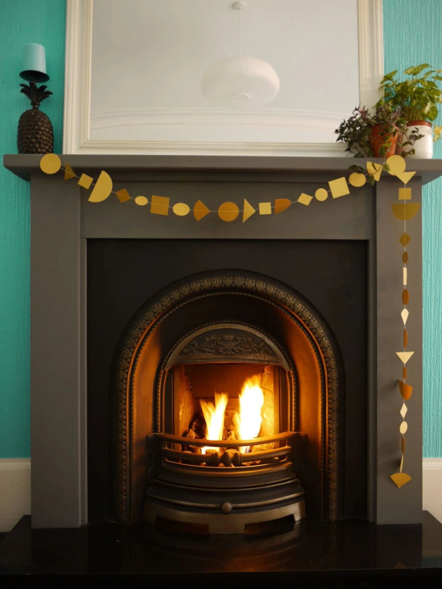 A gold geometric paper garland made of circles, squares, and triangles hangs across a dark fireplace mantel, with a mirror and potted plants above.