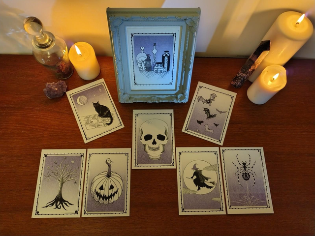 Set of tarot style riso prints with Halloween-themed illustrations on a wooden surface with candles and a framed picture.
