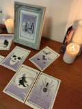 Set of tarot style riso prints with Halloween-themed illustrations on a wooden surface with candles and a framed picture.