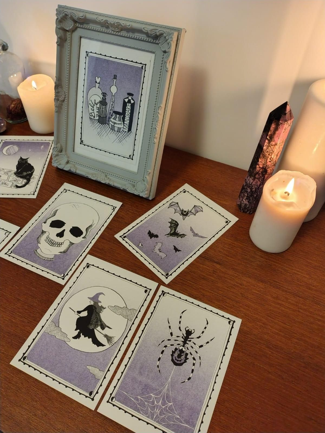 Set of tarot style riso prints with Halloween-themed illustrations on a wooden surface with candles and a framed picture.